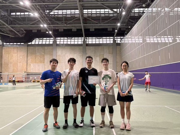 Five team members with a certificate and shuttlecocks on an indoor badminton court.