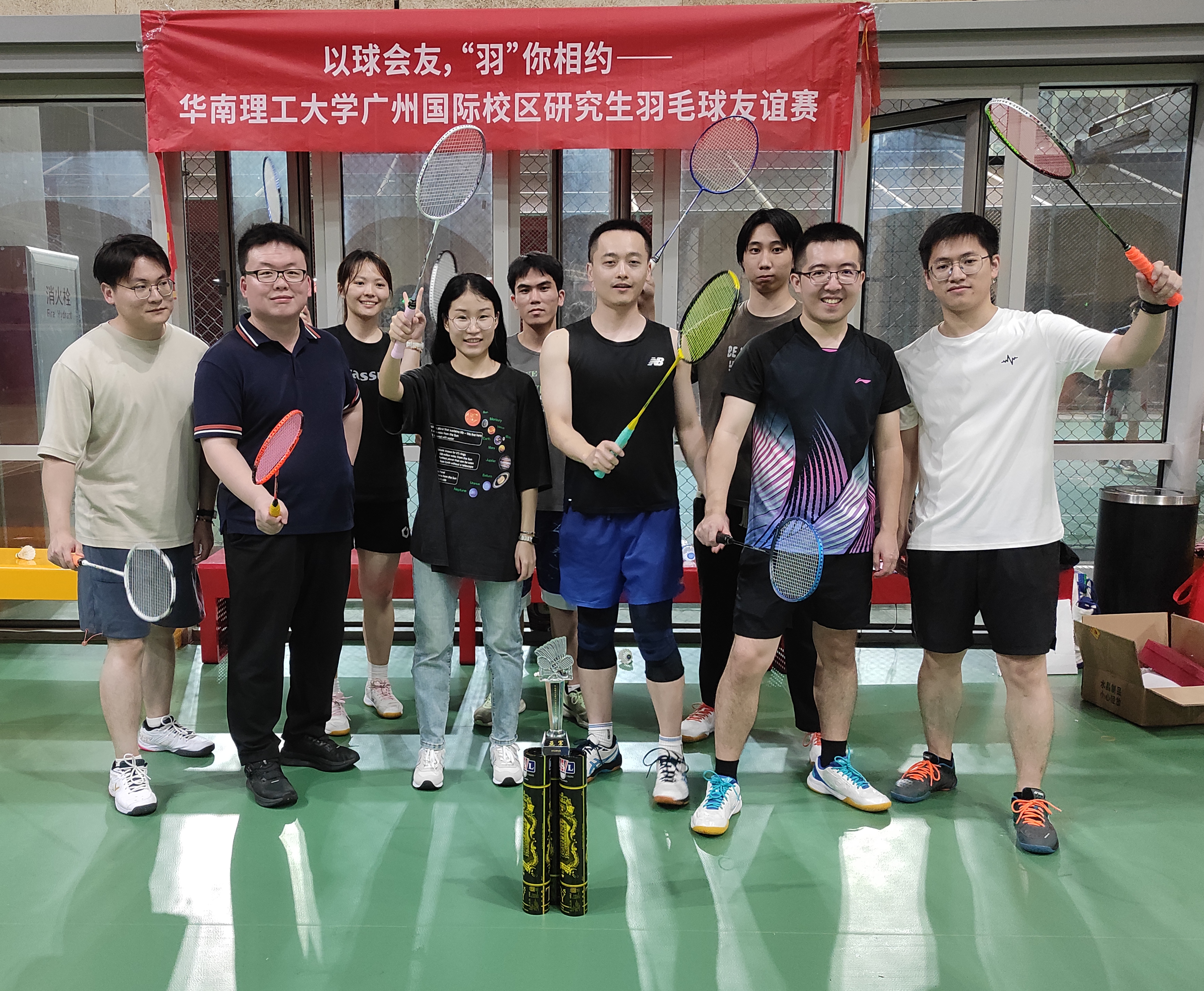 Badminton participants holding rackets on an indoor court beneath the event banner.
