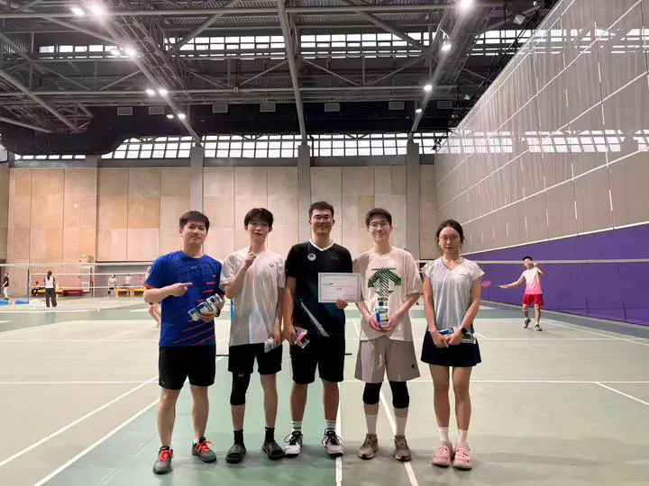 Five team members with a certificate and shuttlecocks on an indoor badminton court.